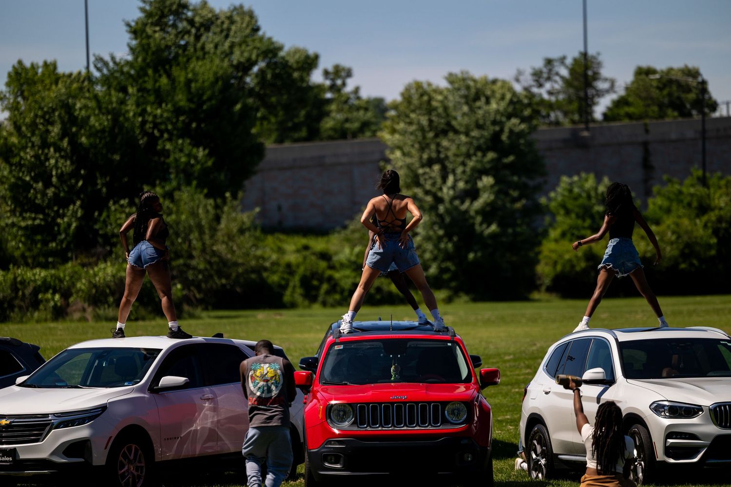 Women dancing on top of cars at an outdoor event on a sunny day with green trees in the background.