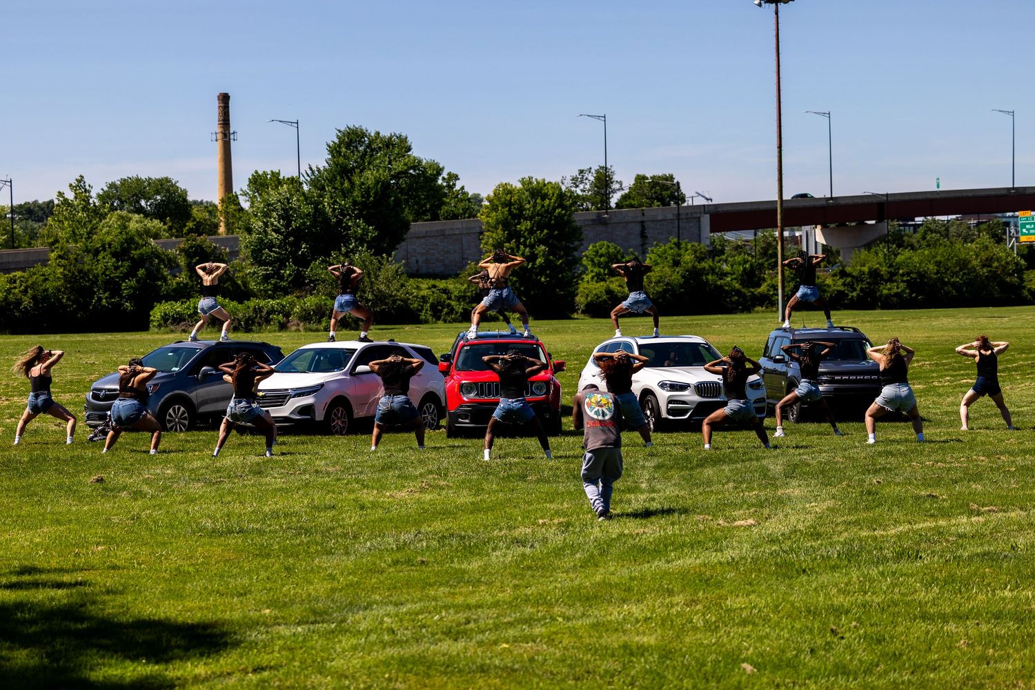 Group of people striking yoga poses on grass and car rooftops outdoors on a sunny day.