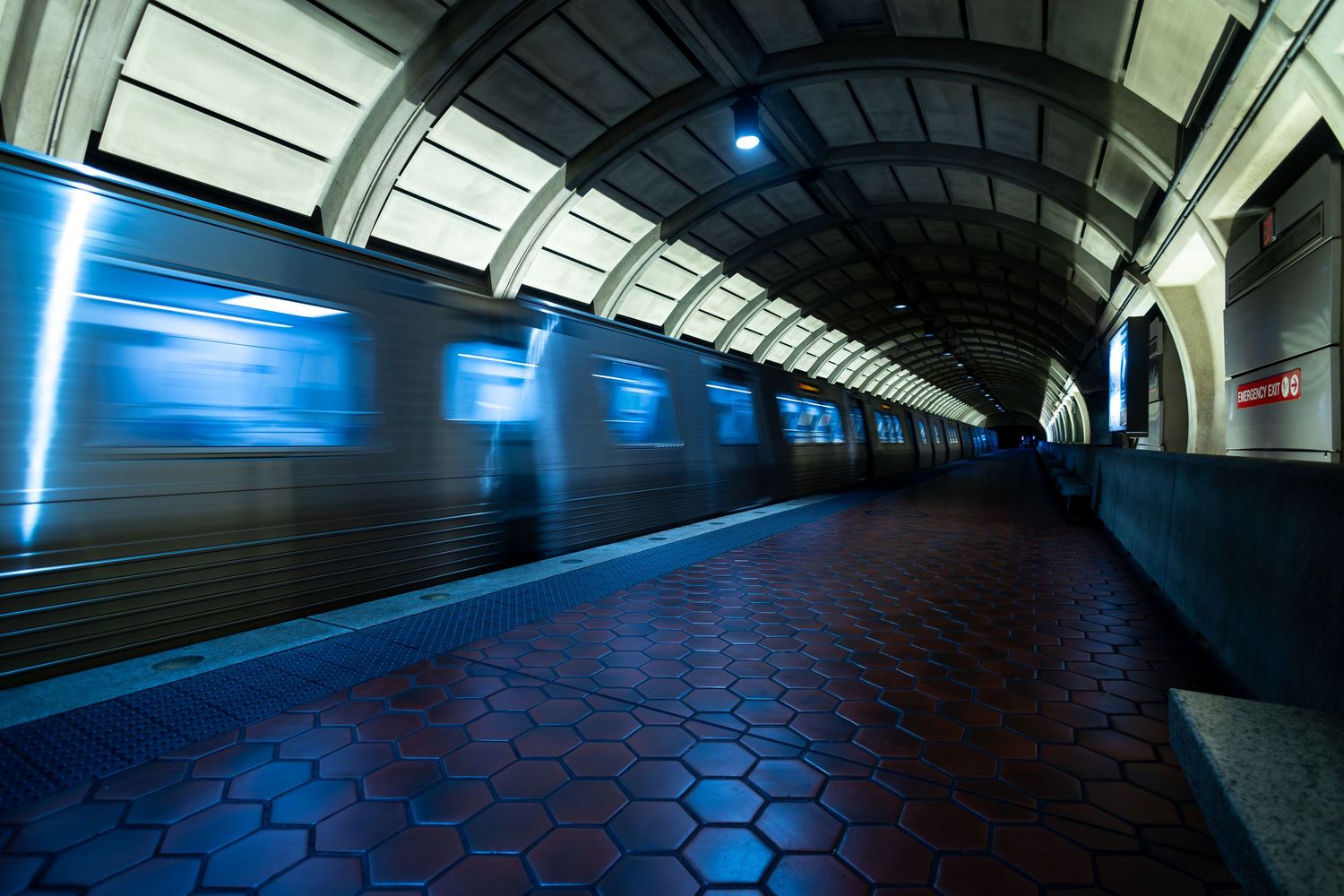 A blurred subway train creates motion streaks of blue light along a curved platform with hexagonal floor tiles in a metro station.