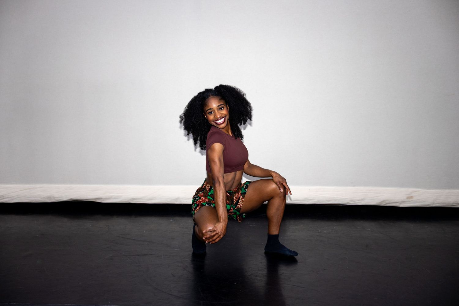 A joyful dancer in colorful African print skirt smiles while posing in a low squat on stage.