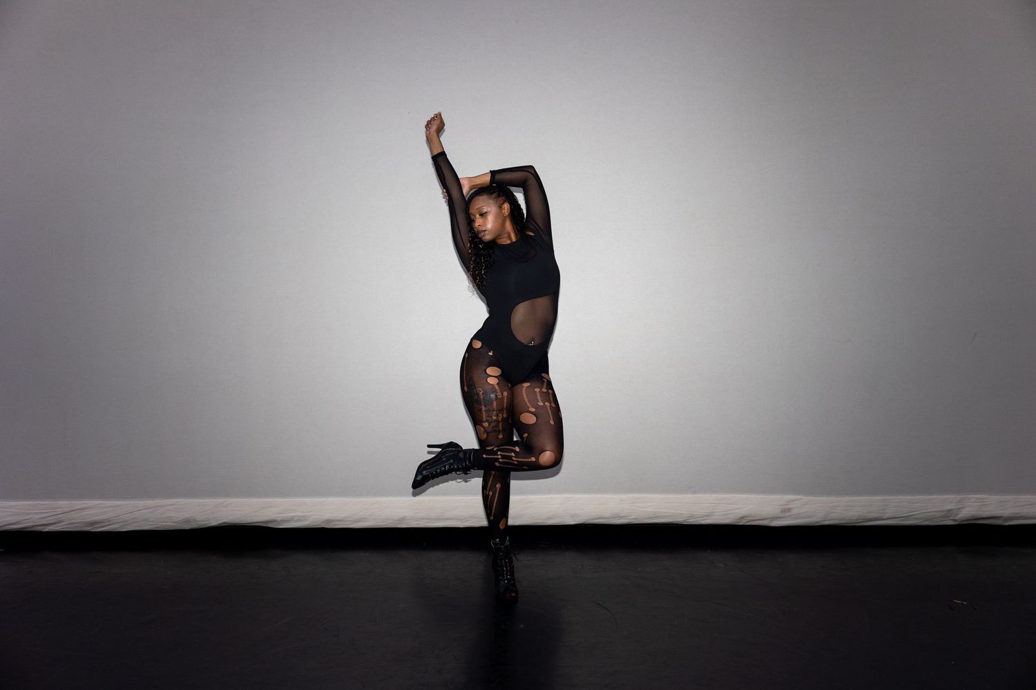 A dancer in black mesh bodysuit performs a dramatic one-legged balance with arm raised overhead.