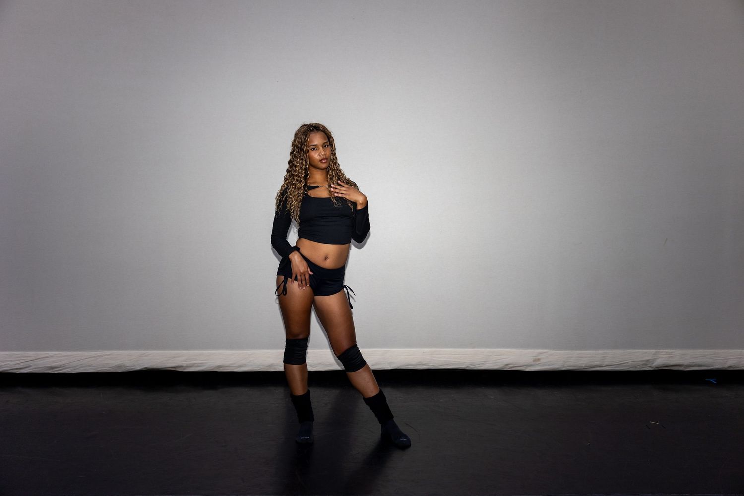 A woman in black crop top, shorts, and knee-high boots strikes a confident pose against gray backdrop.