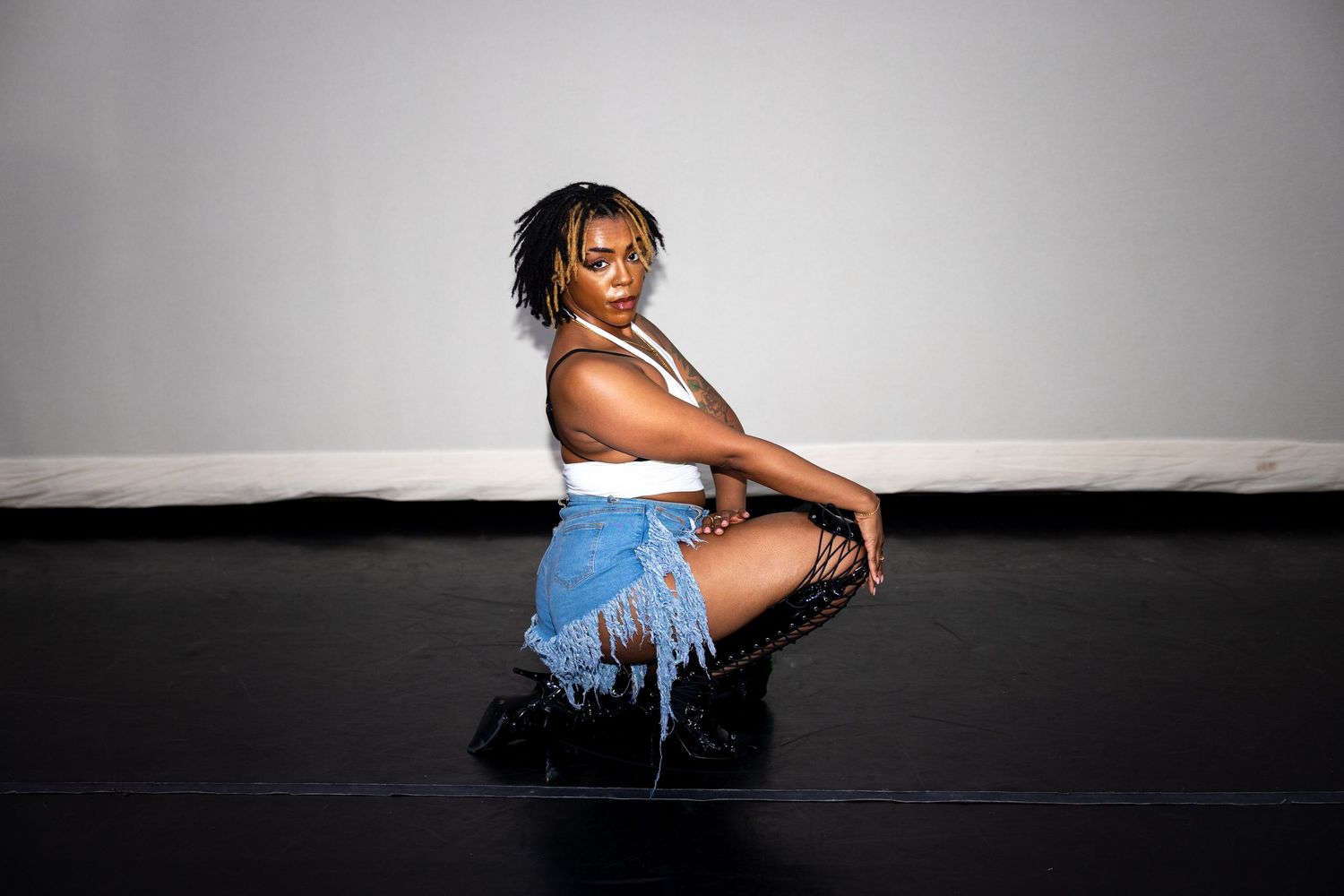Woman in white crop top and frayed denim shorts kneeling on black studio floor.