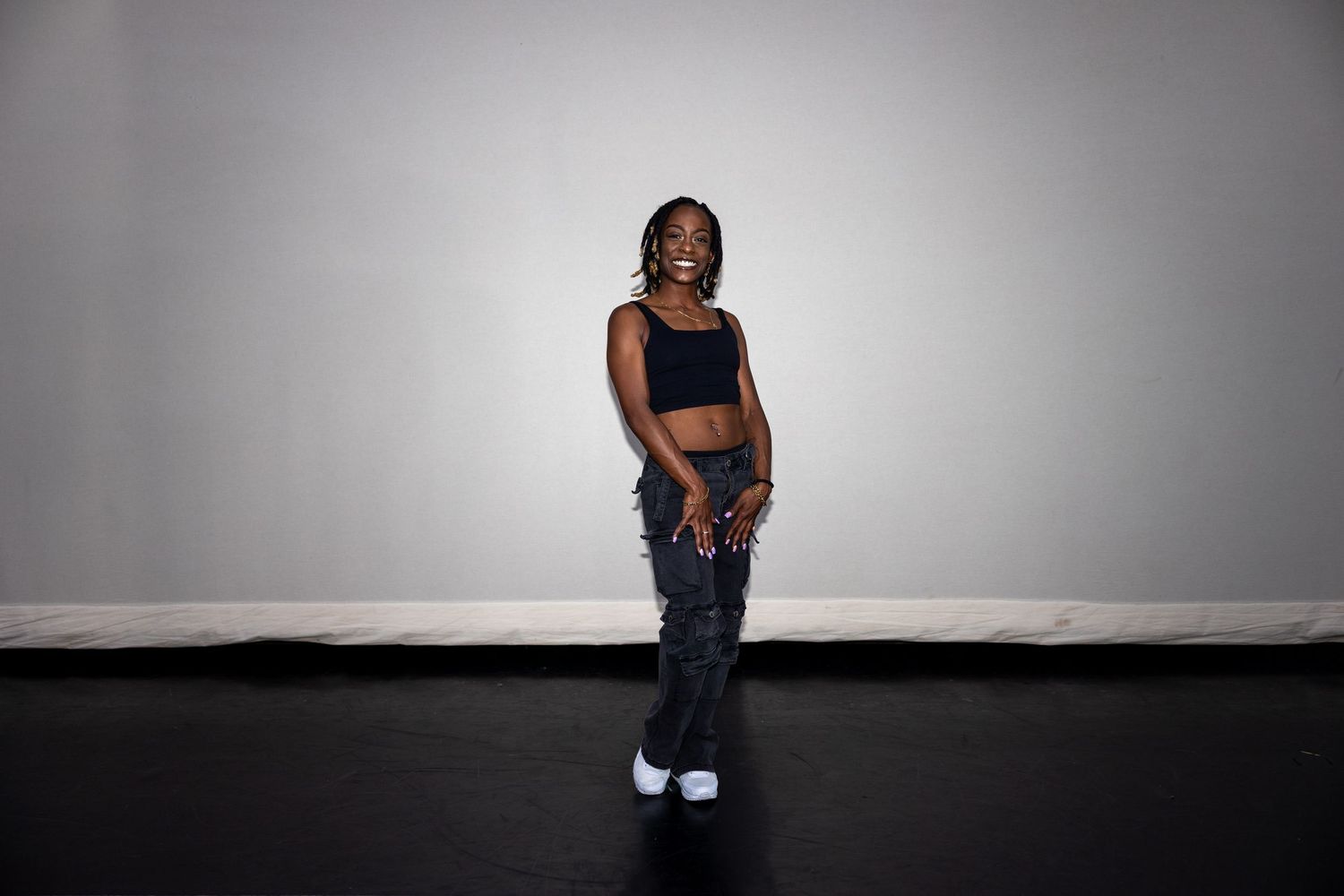 Smiling Black woman in black crop top and cargo pants stands on dark studio floor.