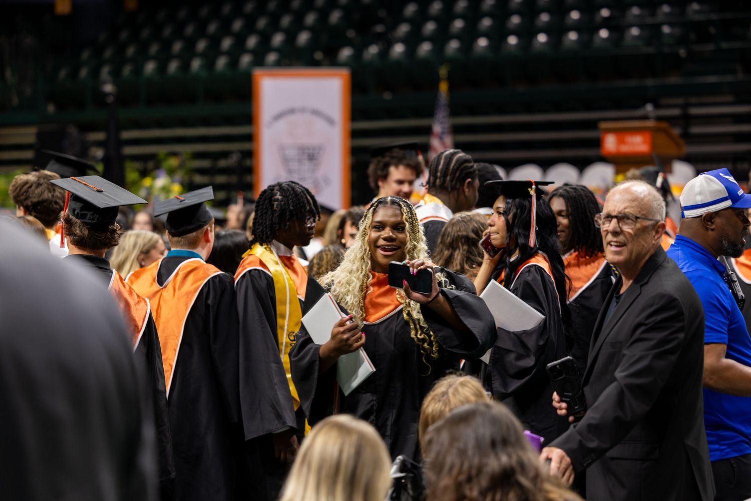 College graduates in black and orange robes celebrate at an indoor graduation ceremony in an arena.