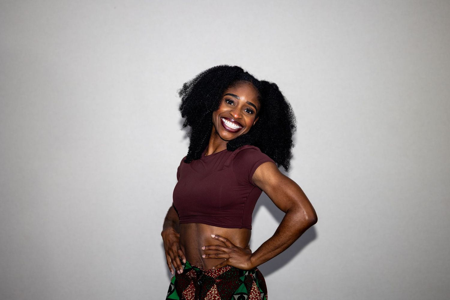 Smiling Black woman with natural afro hair wearing a maroon crop top and printed skirt.