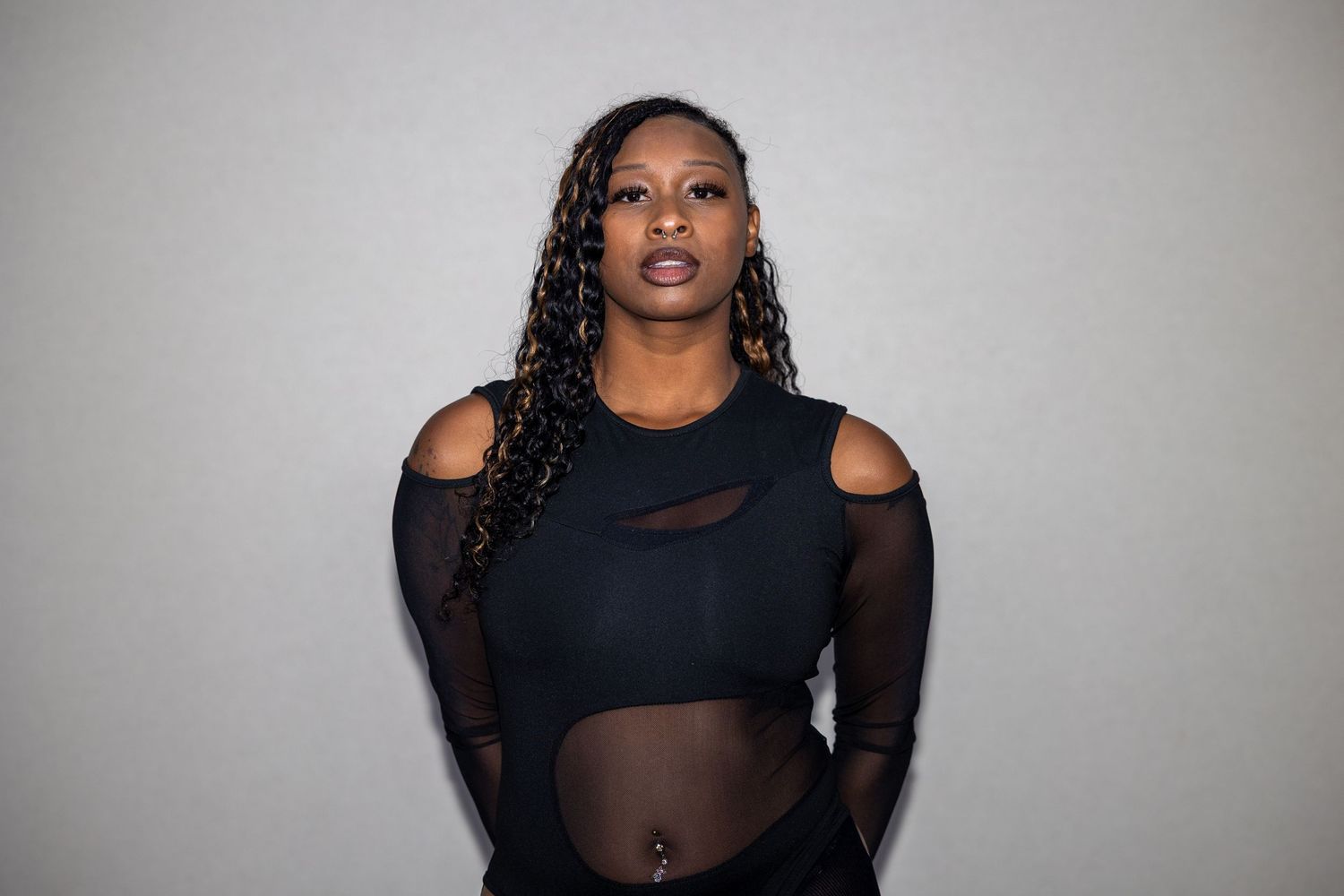 A confident Black woman poses in a black cutout mesh top and pants against a light gray background.