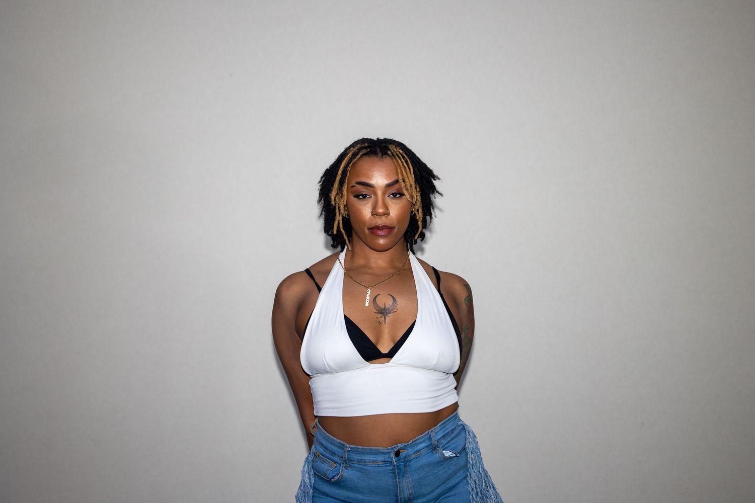 Confident Black woman in white halter crop top and denim jeans poses against a gray background.