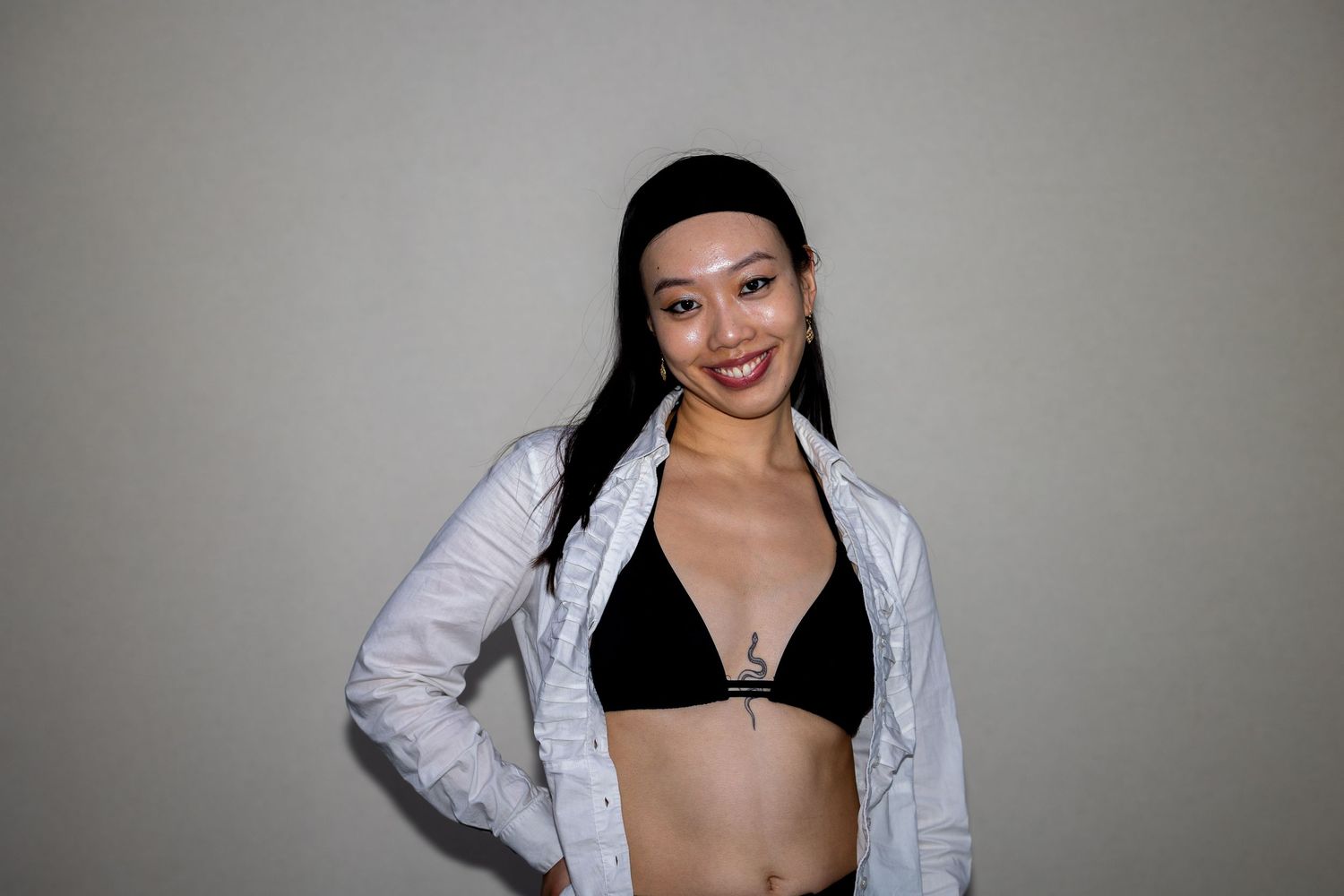 Smiling Asian woman in black bikini top and open white shirt posing against gray background.