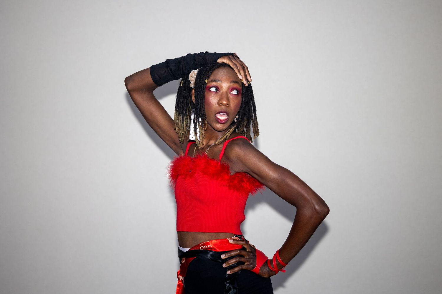 A Black woman poses confidently in a red feathered crop top, black gloves, and patterned pants against a gray background.