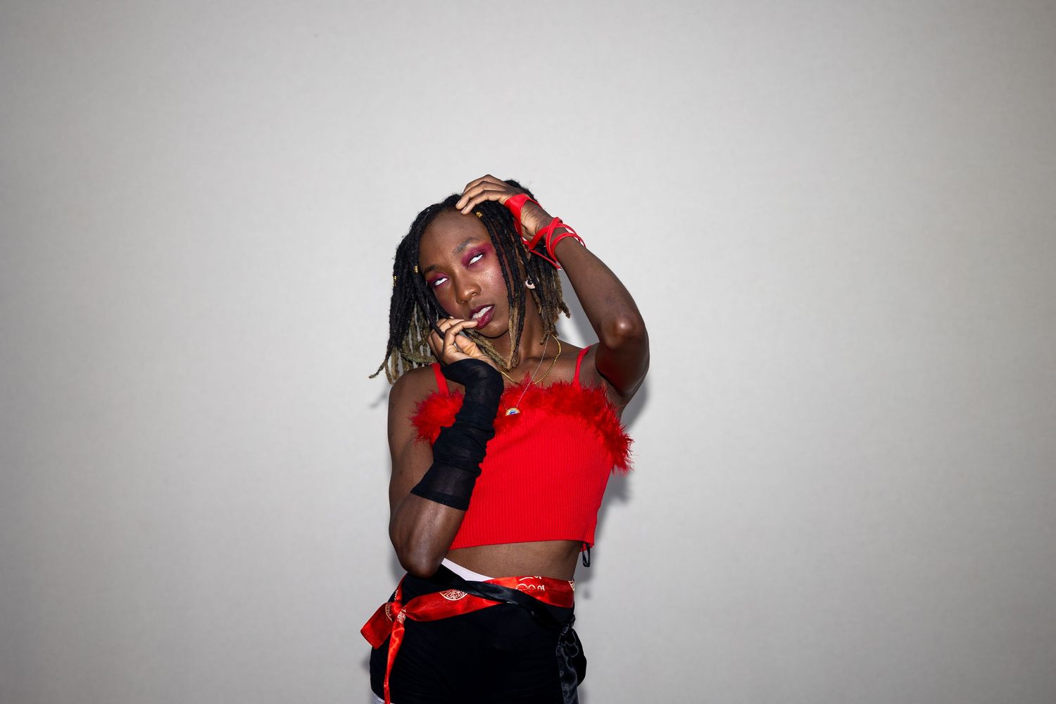 Confident performer in red feathered crop top with black gloves posing thoughtfully against a neutral background.