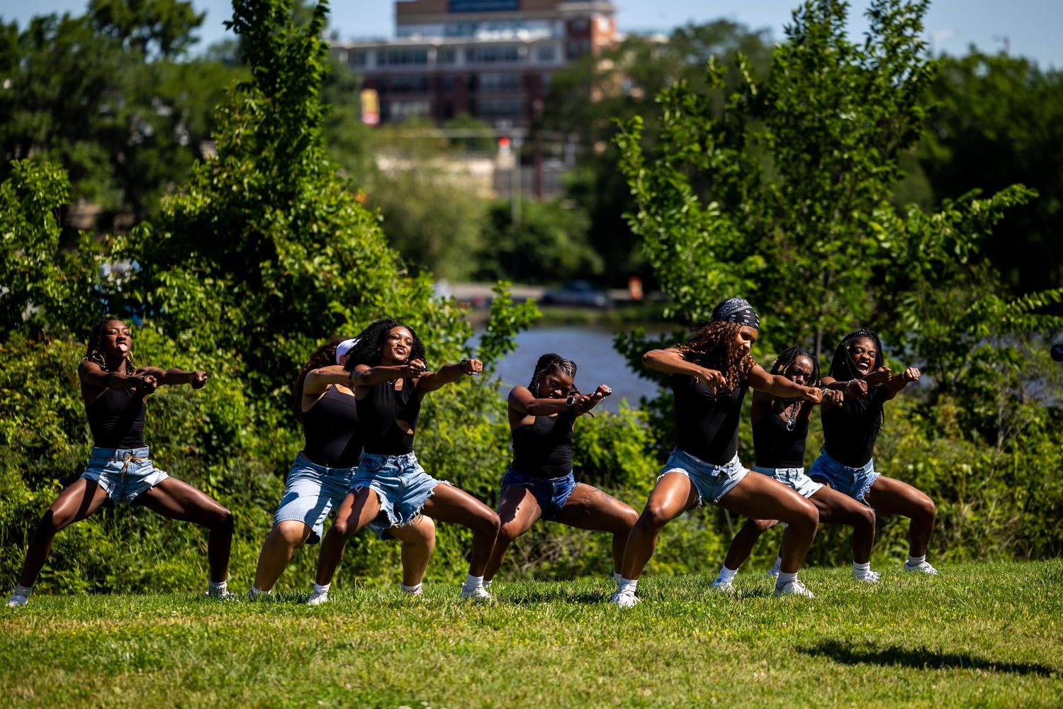 Group of dancers performing synchronized moves outdoors on grass with trees and building in background on sunny day.