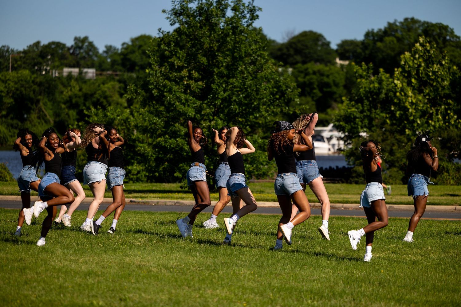 A group of young women in black tops and denim shorts dancing outdoors in a park near a lake.