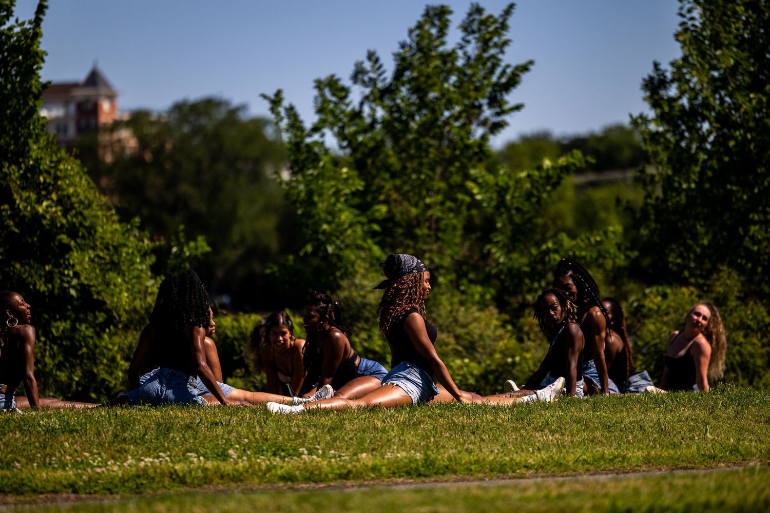 Group of young women relaxing on green grass in a sunny park surrounded by lush trees on a bright summer day.