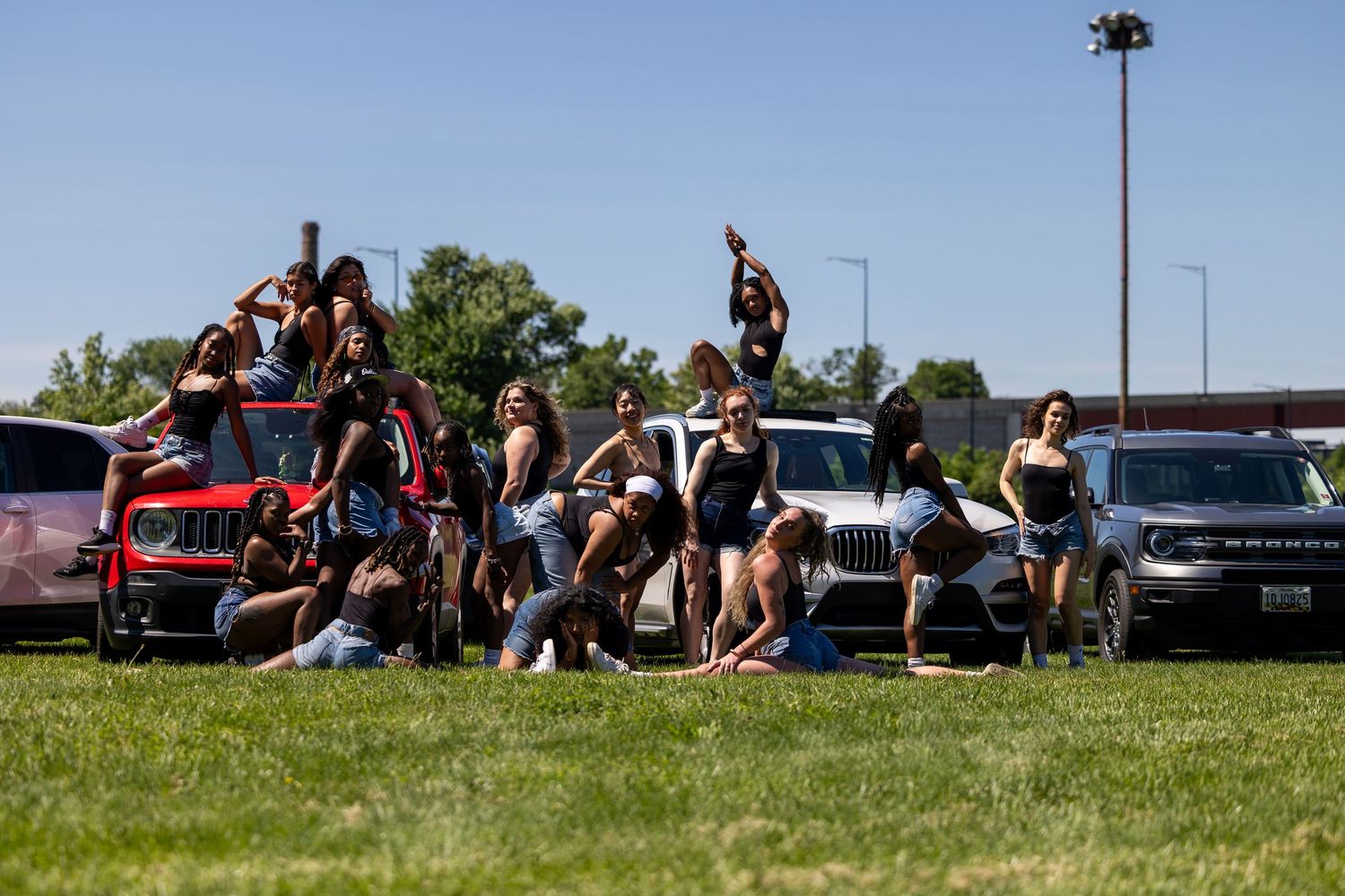 Group of young women posing outdoors on grass near parked SUVs on a sunny day.