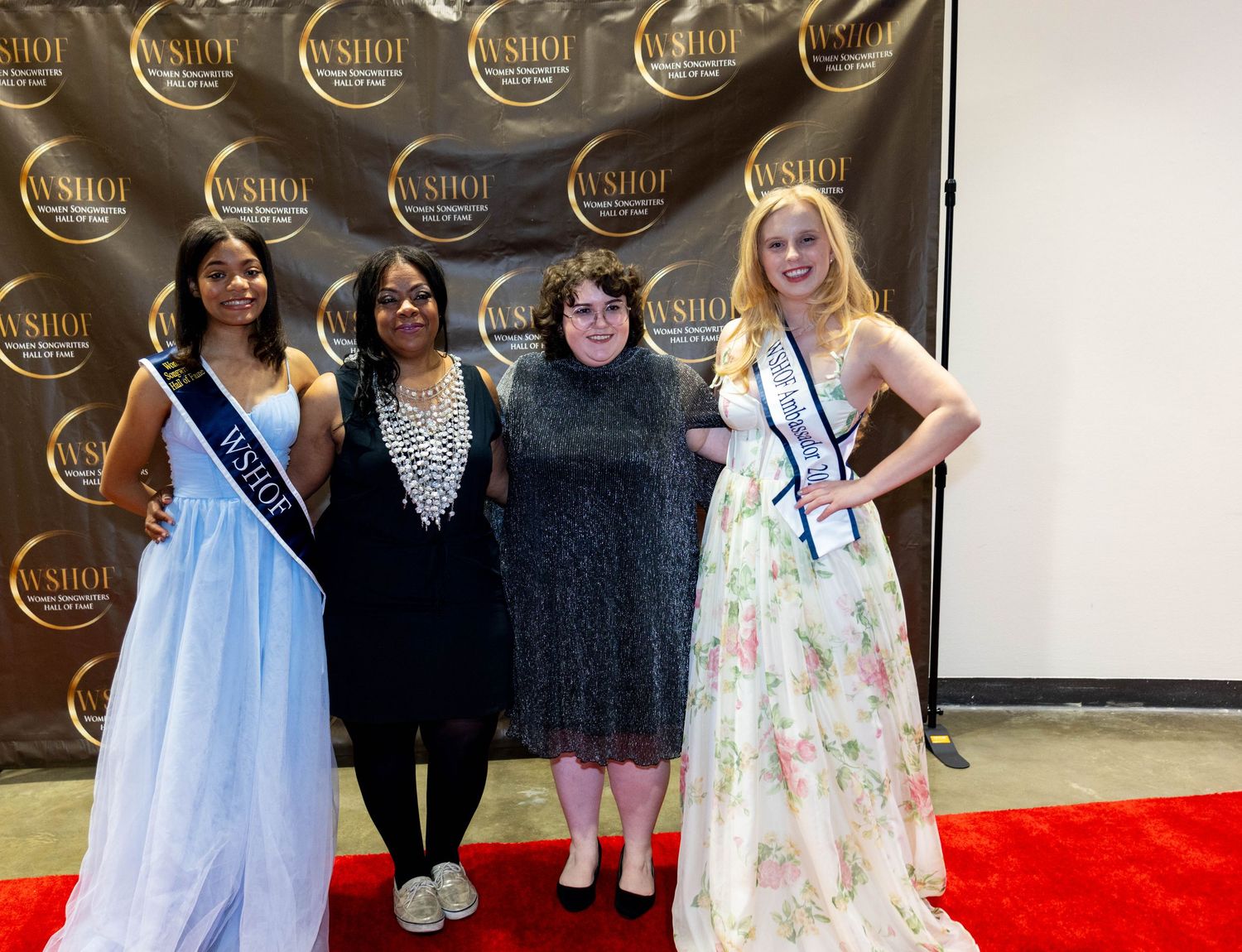 Four women pose on red carpet at WSHOF Women Songwriters Hall of Fame event, two wearing pageant sashes and formal gowns.