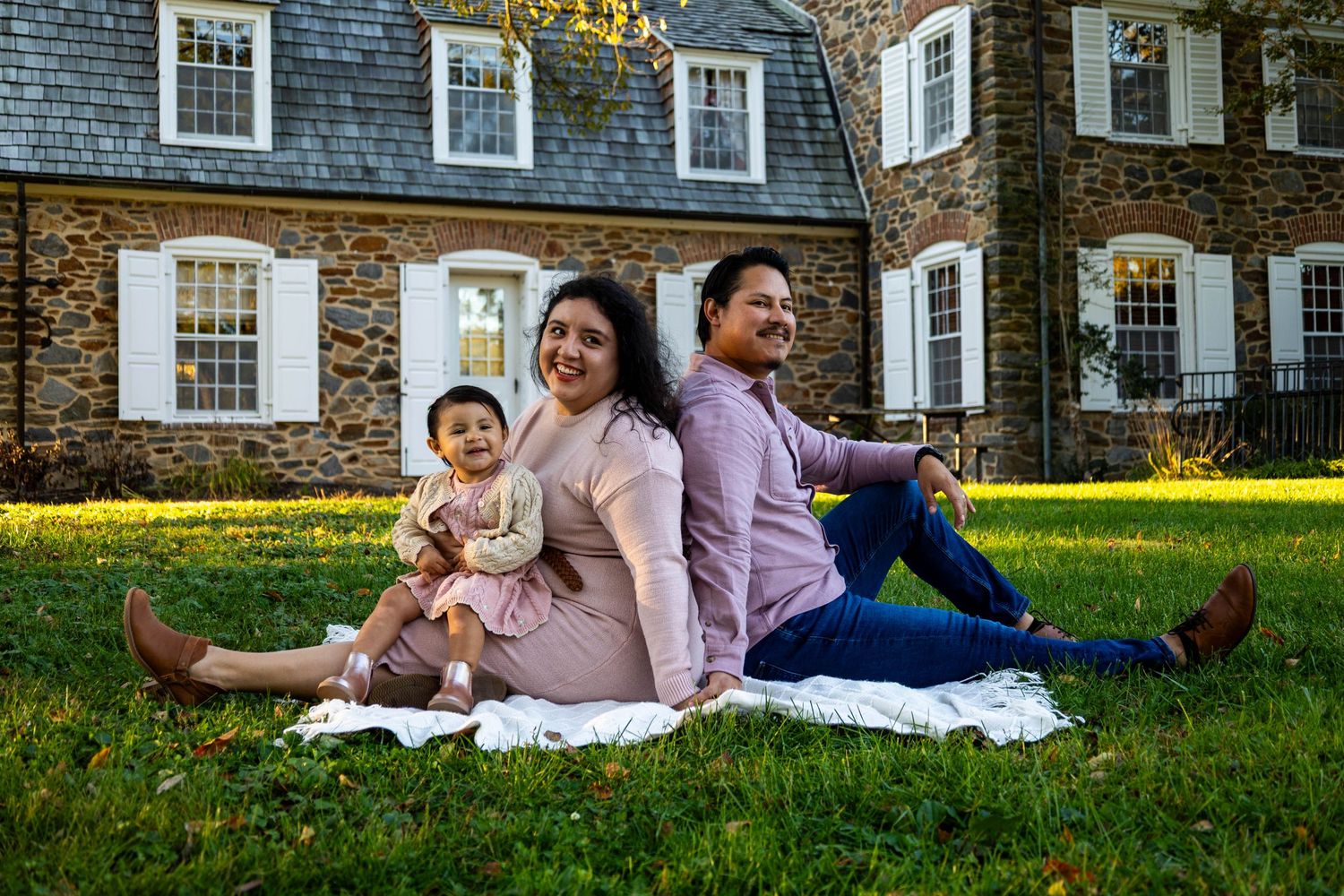 Family of three sitting on a blanket on green grass in front of a historic stone building on a sunny day.