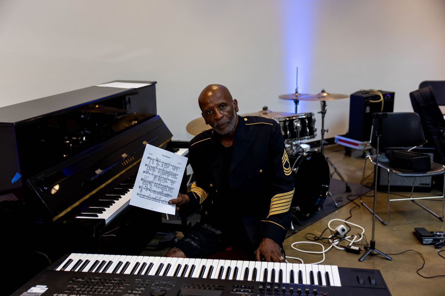 Musician in military uniform holds sheet music beside a grand piano and keyboard in a music studio.