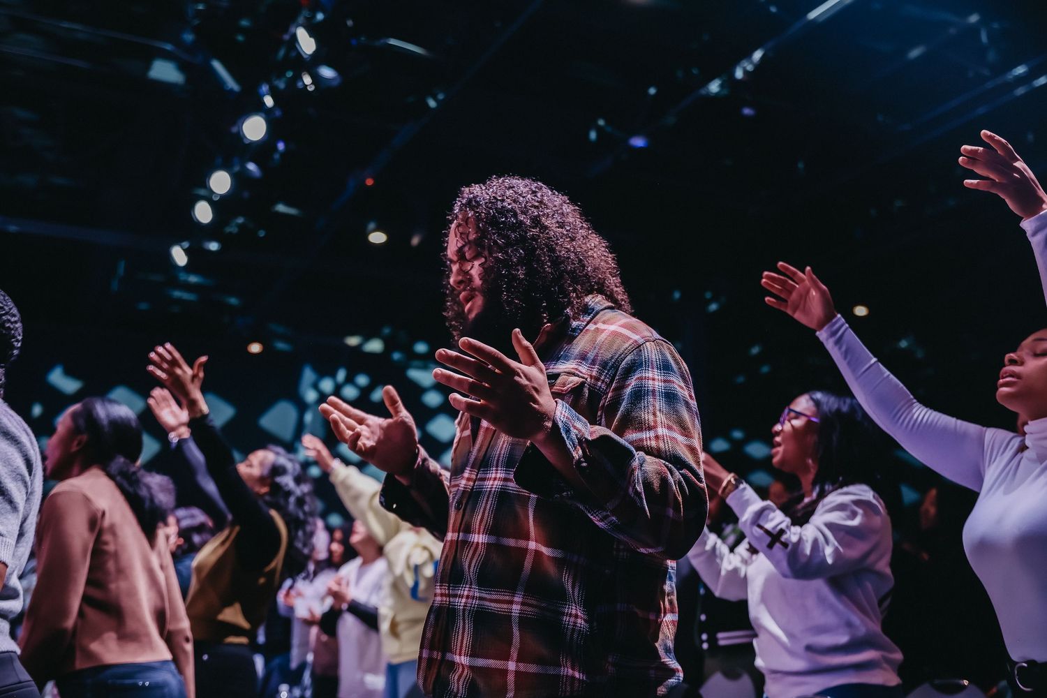 A diverse group of people worshipping with raised hands at a church service in a dimly lit venue.