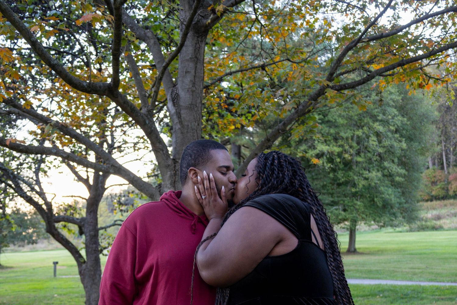 Couple sharing a romantic kiss outdoors in a lush green park with colorful autumn trees in the background.