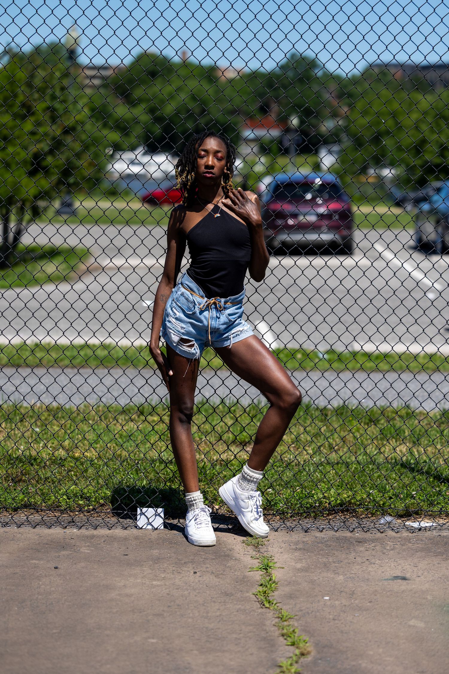 Young Black woman in black bodysuit and denim cutoffs poses by chain-link fence outdoors on sunny day.