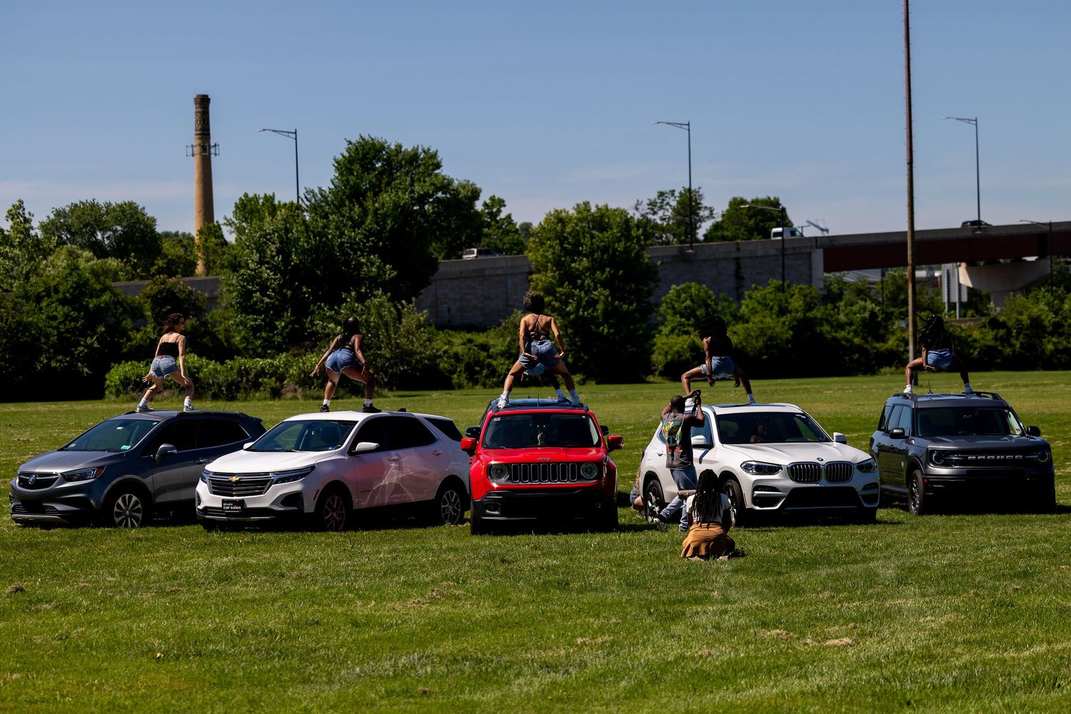 Acrobats perform stunts on top of parked SUVs and cars on a green field with industrial buildings in background.