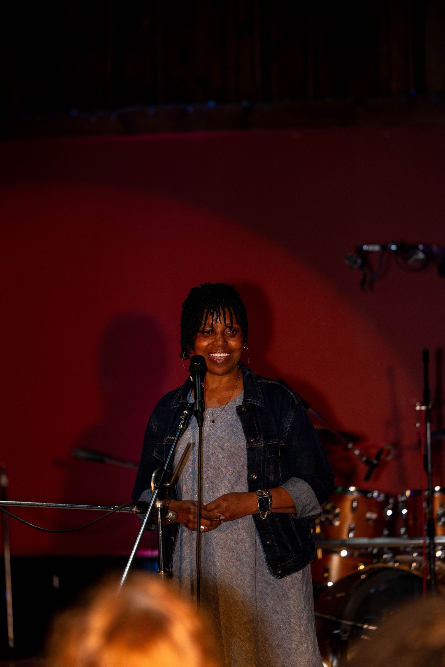 Woman smiling at microphone on stage with drum kit behind her in red-lit venue.