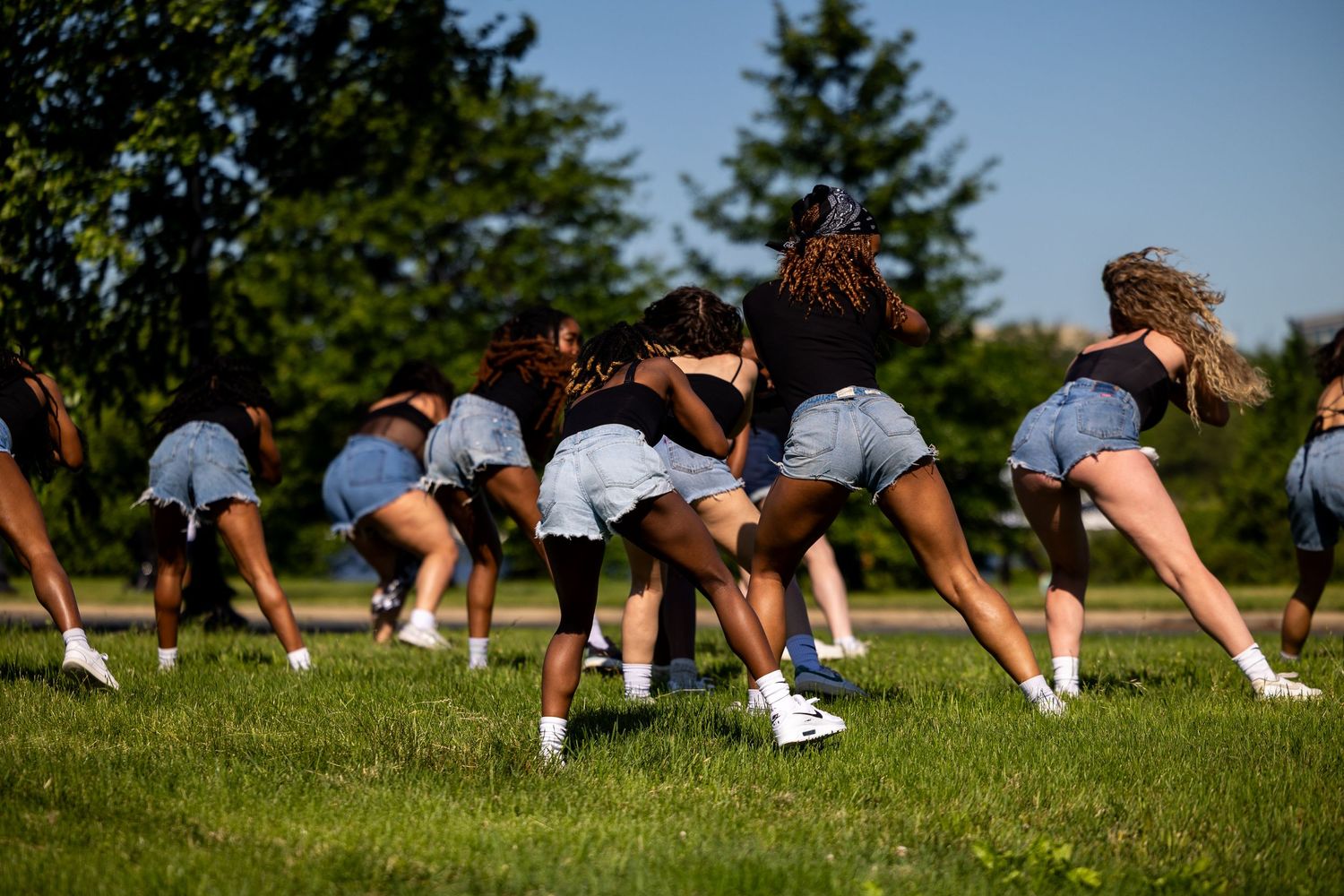 Group of young women in denim shorts performing a synchronized dance routine outdoors on a green lawn.