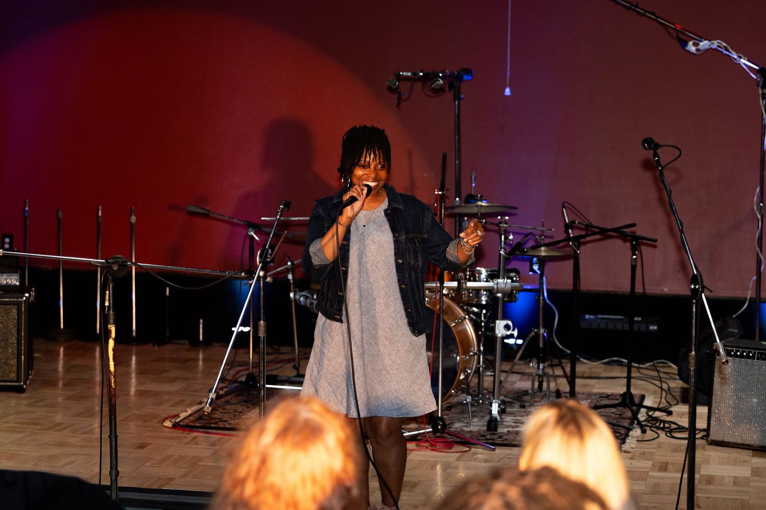 A female singer performs on stage with a microphone, backed by a drum kit in a warmly lit venue.