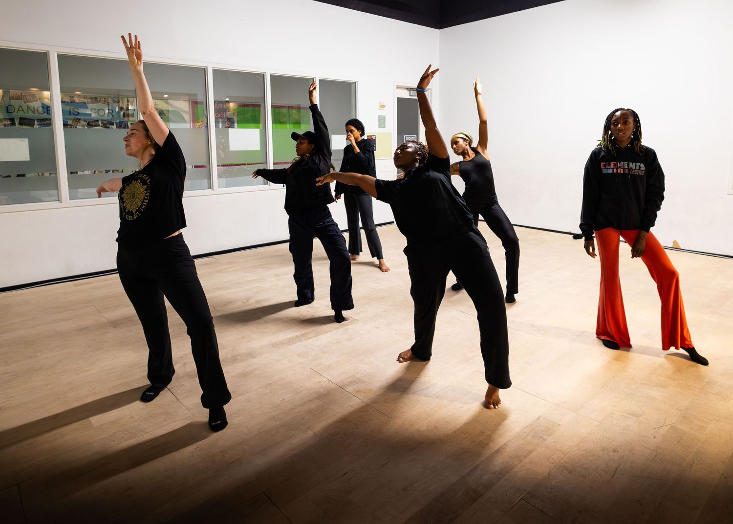 Dancers in black attire perform expressive modern dance moves in a bright studio with wood floors.