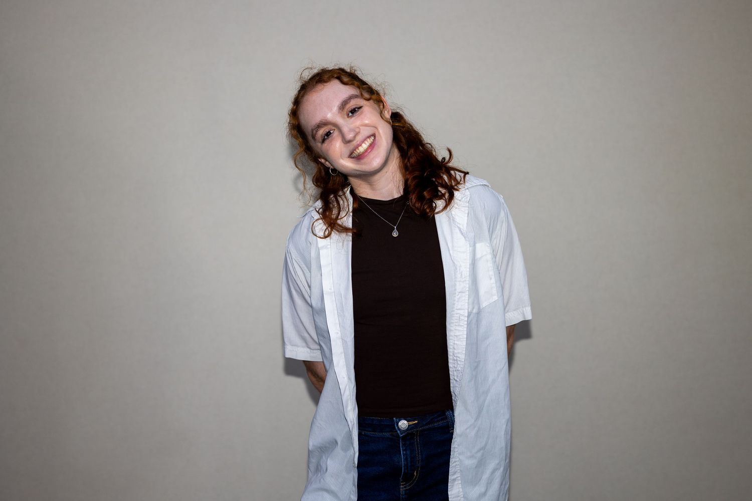 Young woman with curly red hair smiling in black tee and white open shirt with jeans against neutral background.