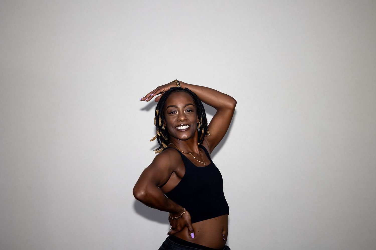 Joyful Black woman in black sports bra dancing with arms raised against white background.