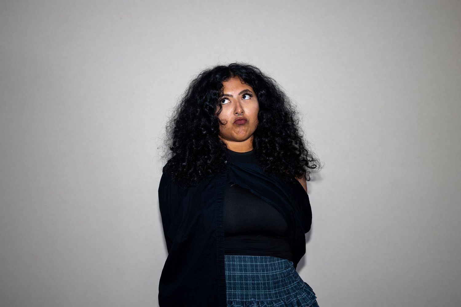 Woman with voluminous curly black hair in black top and plaid skirt looking upward against gray background.