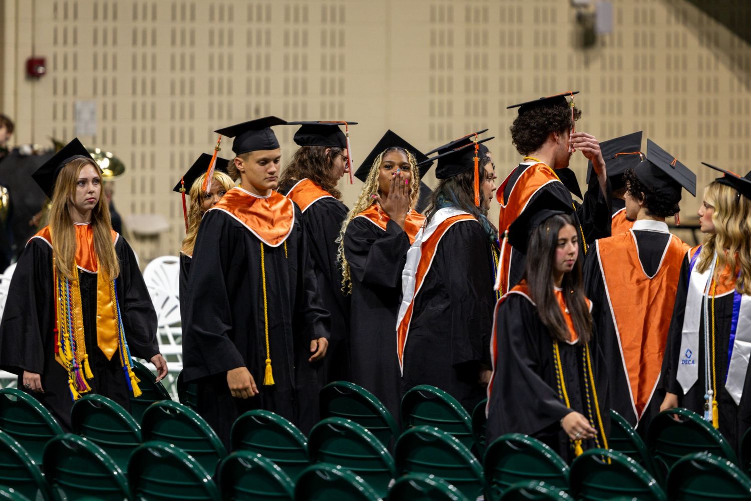 Students in black and orange graduation gowns and caps stand in rows at a high school commencement ceremony.