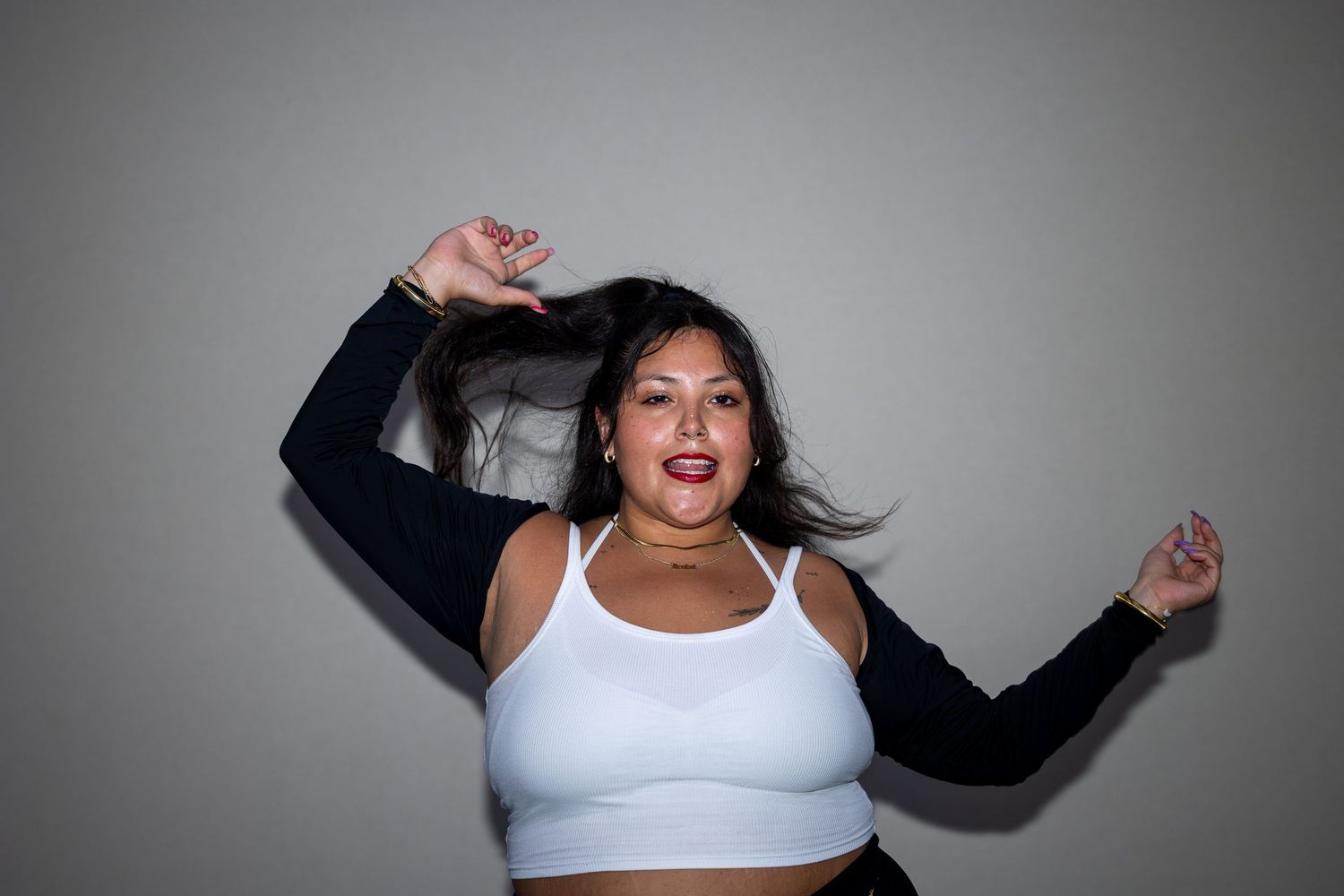 Woman in white crop top and black sleeves dancing energetically with hair flowing against gray background.