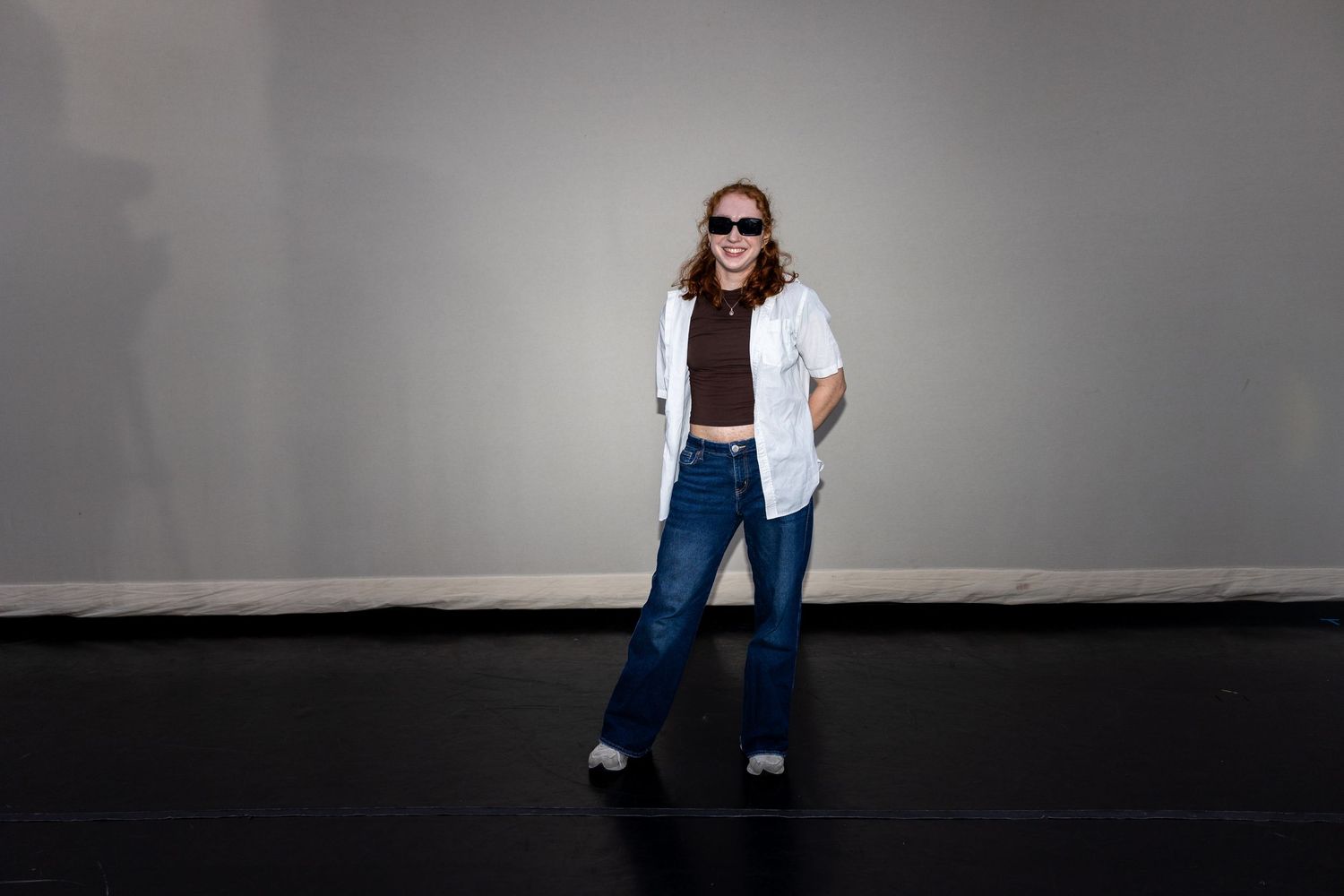 A performer in white shirt, jeans, and sunglasses laughs while posing on a black stage floor.