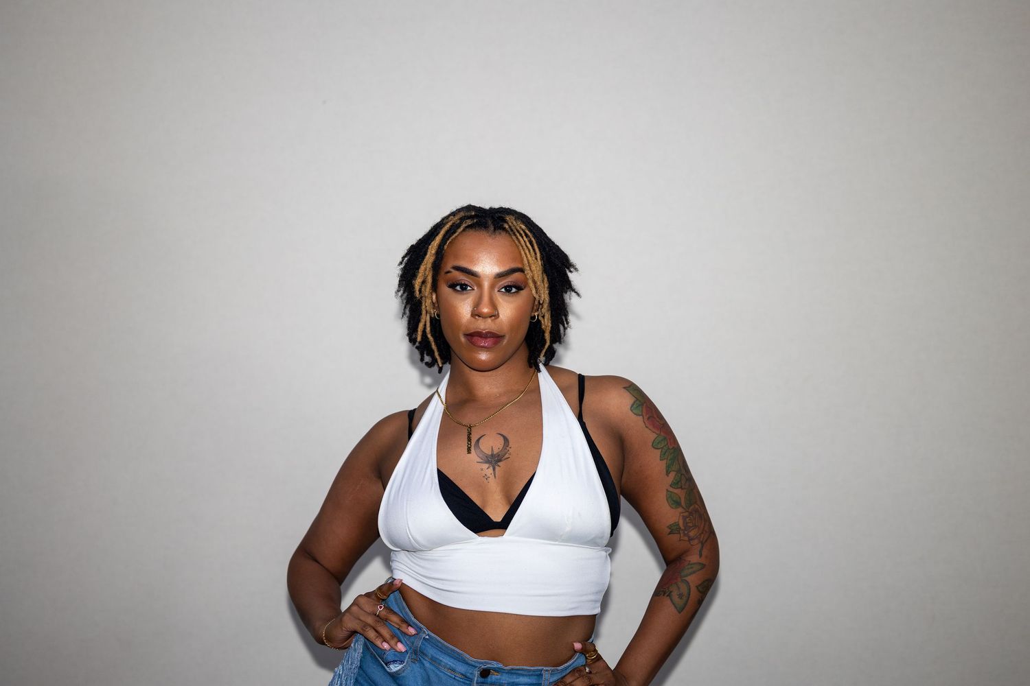 Young woman with braids and tattoos posing in a white halter crop top and jeans, serious expression.