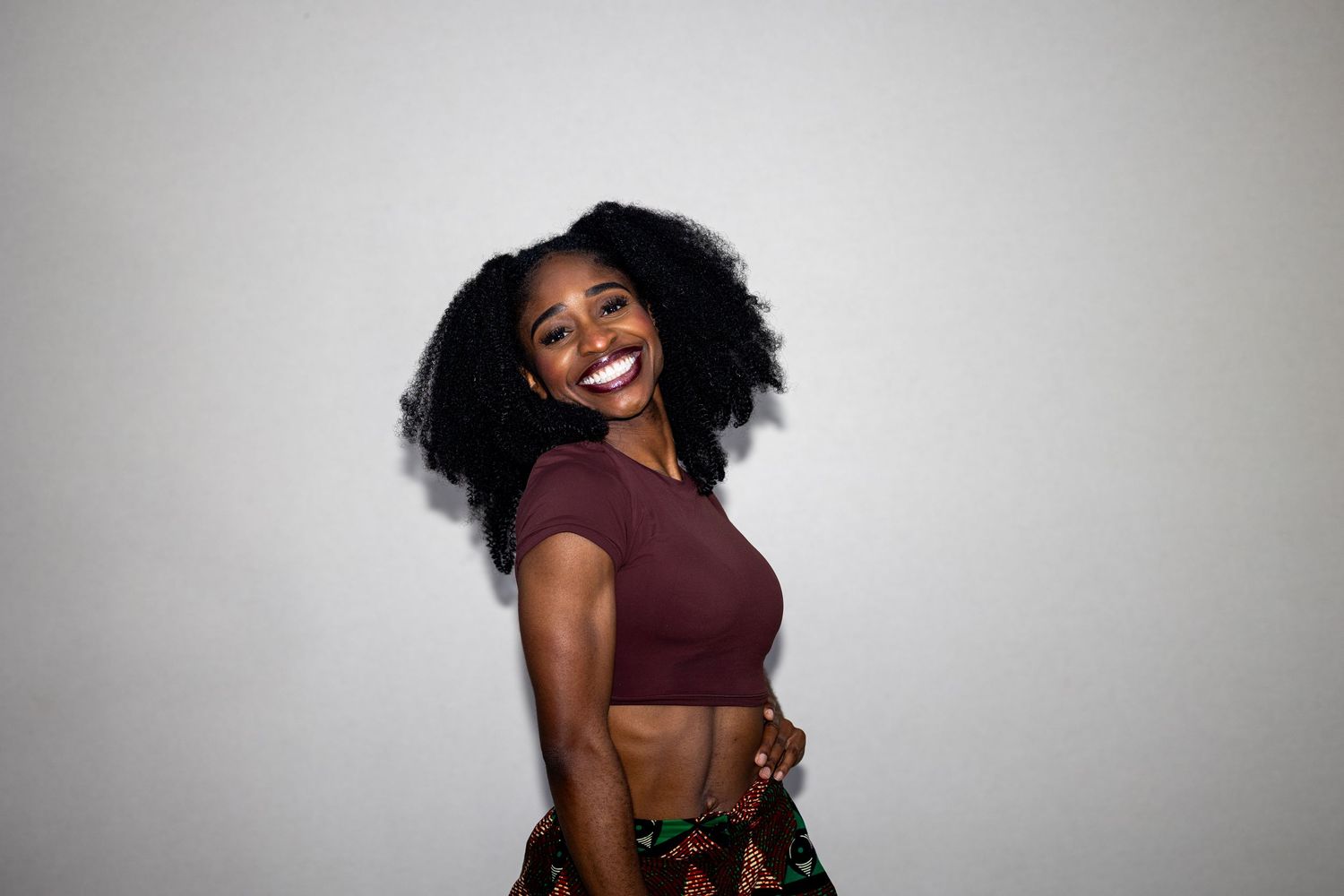 Joyful young Black woman with natural afro hair laughing in a maroon crop top and colorful printed skirt.