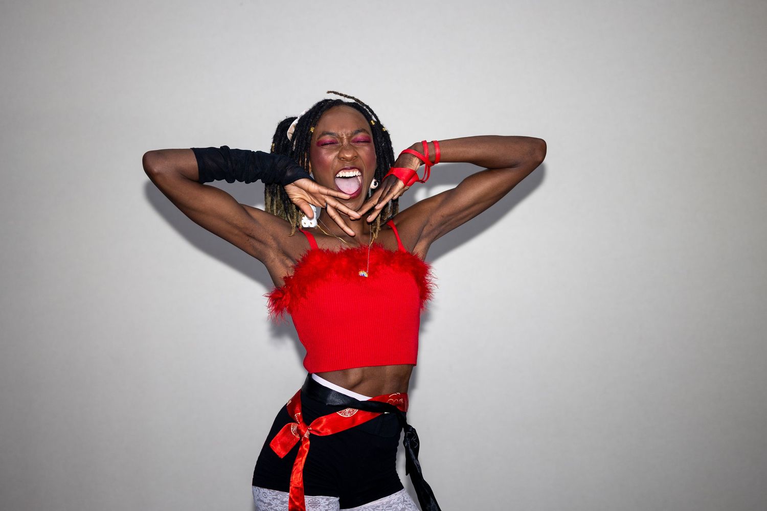 Expressive performer in red feathered crop top and black gloves screaming while adjusting braided hair accessories.