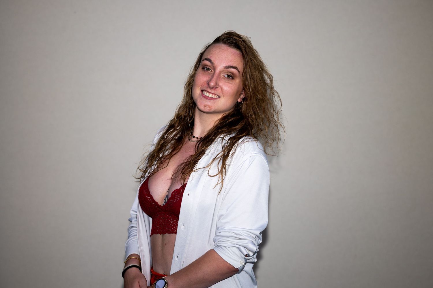 Smiling woman in white blazer and burgundy bralette poses against light wall.