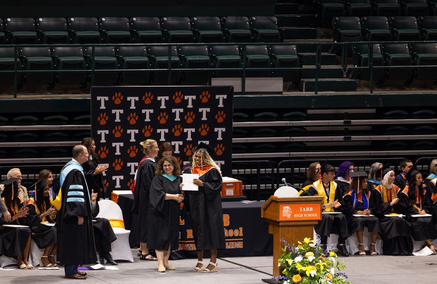 Faculty and graduates in academic regalia shake hands on stage during a university graduation ceremony indoors.