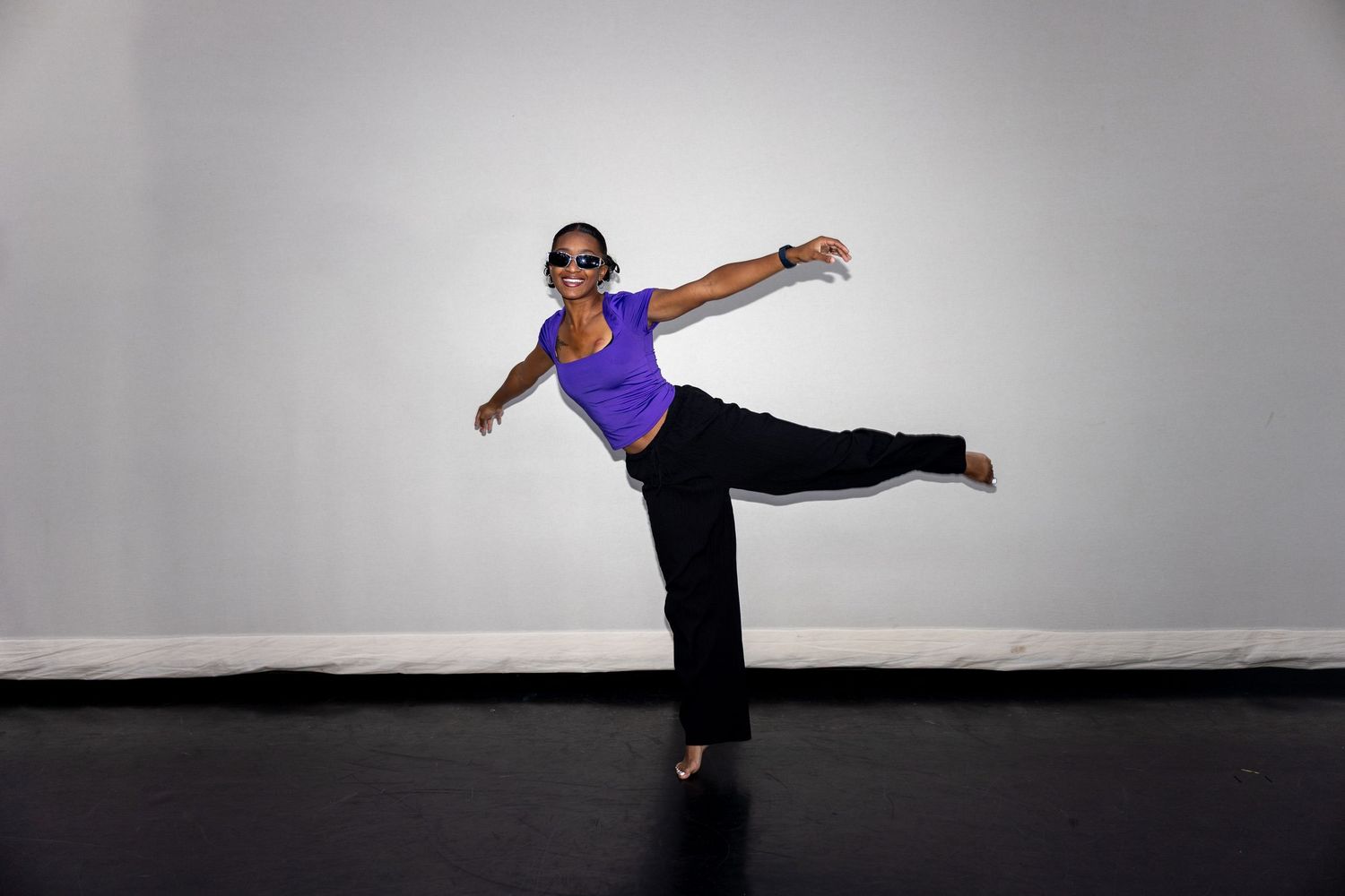 A dancer in a purple top and black pants balances on one leg with arms outstretched in a studio.
