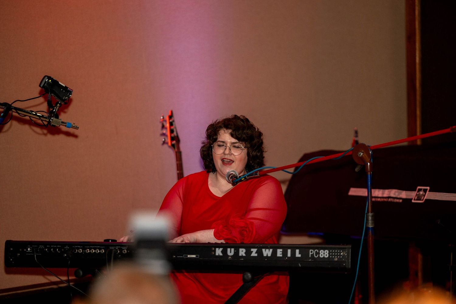 Woman in red dress performing at a Kurzweil keyboard on stage with a guitar visible in the background.