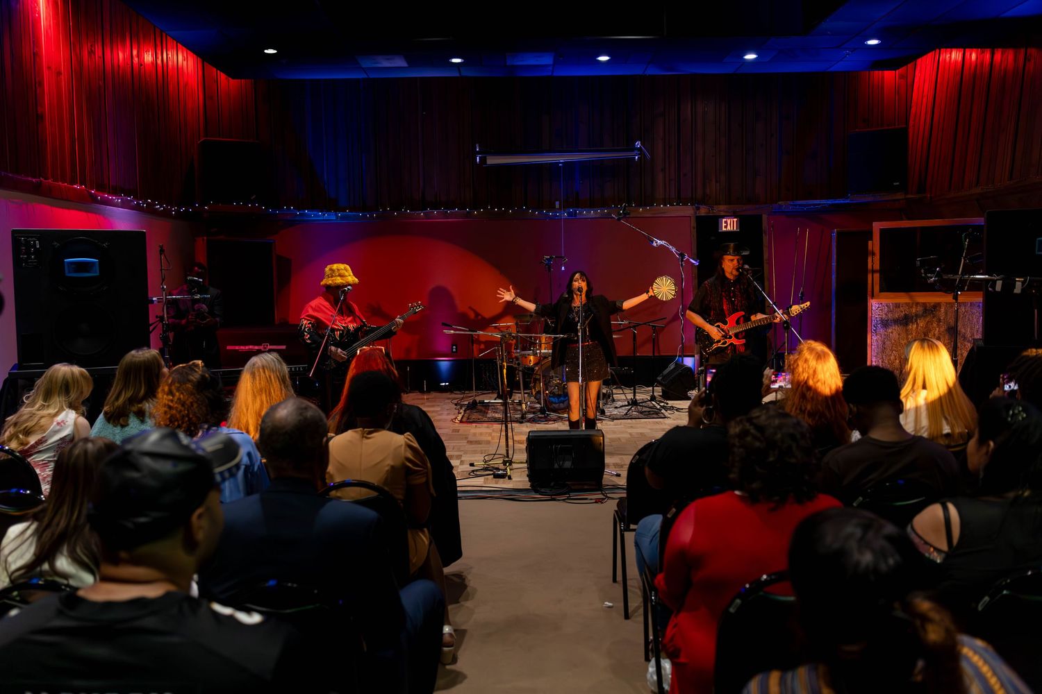 Live band performs on a small red-lit stage for an intimate seated audience in a cozy music venue.