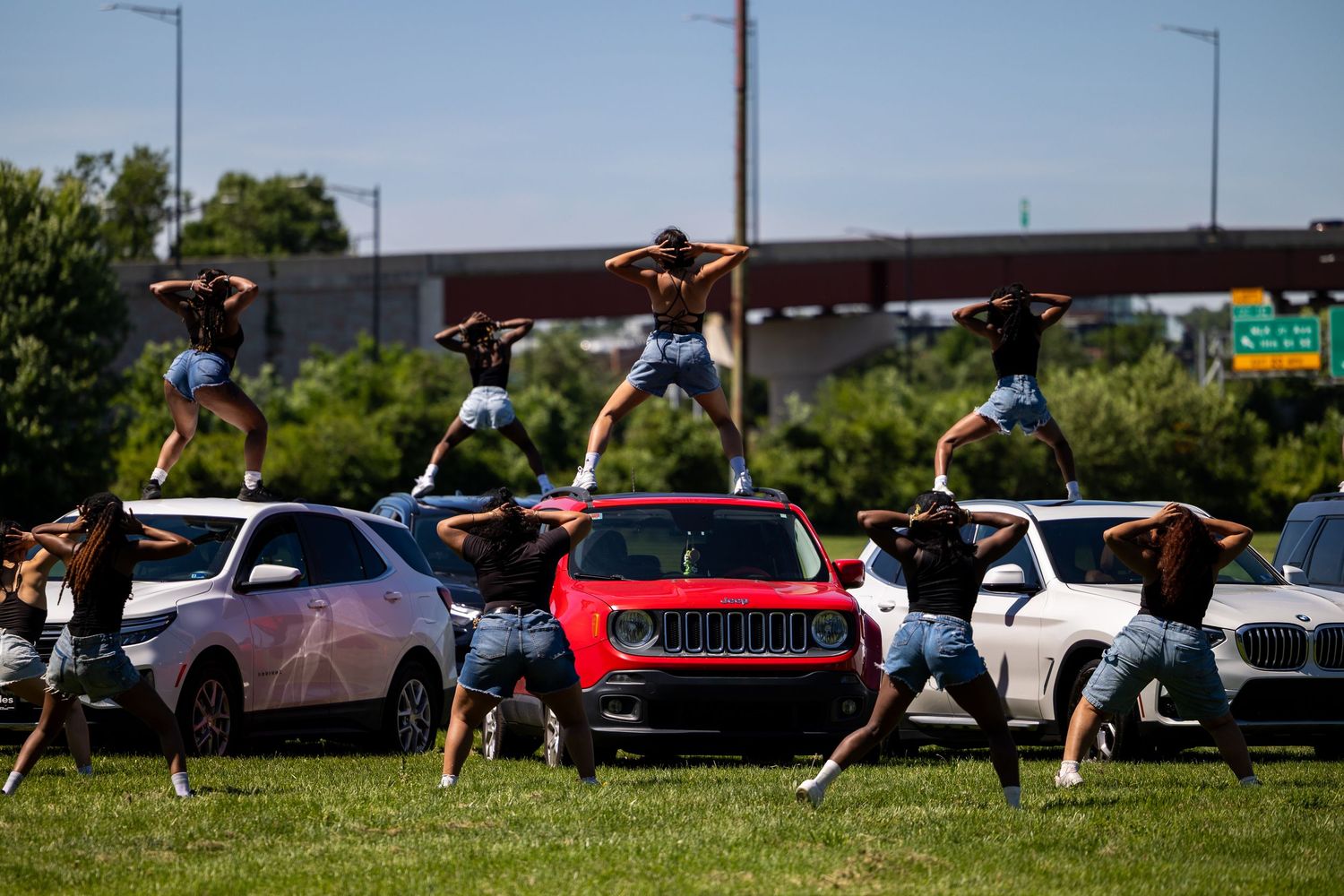 Cheerleaders perform acrobatic stunts on top of cars in a parking lot on a sunny day.