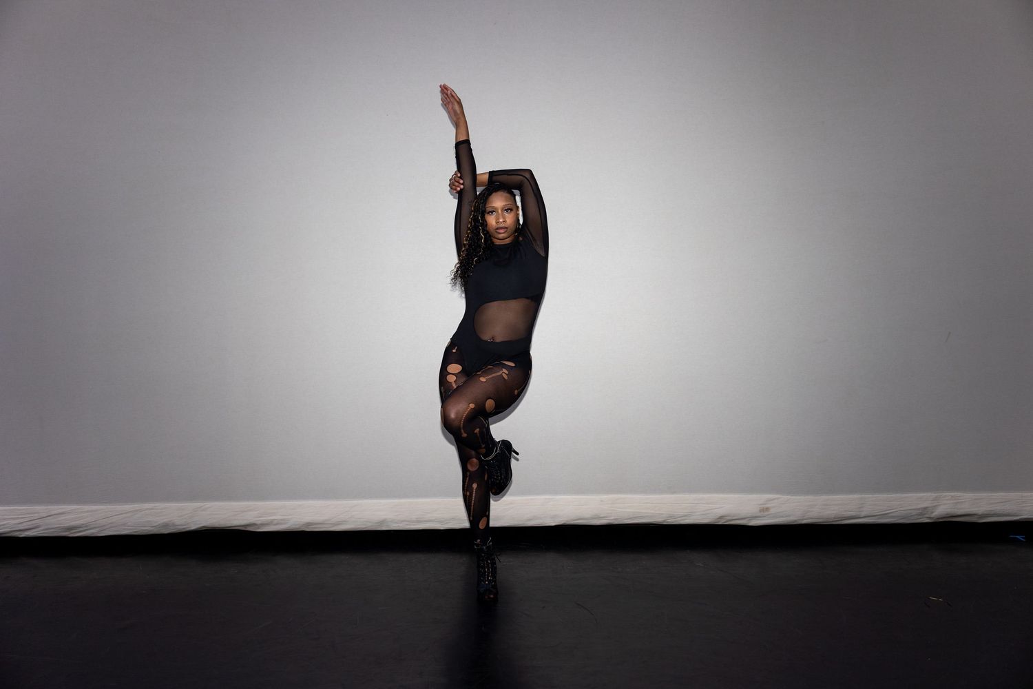 A dancer in black sheer bodysuit executes a graceful one-legged pose with arm extended upward.