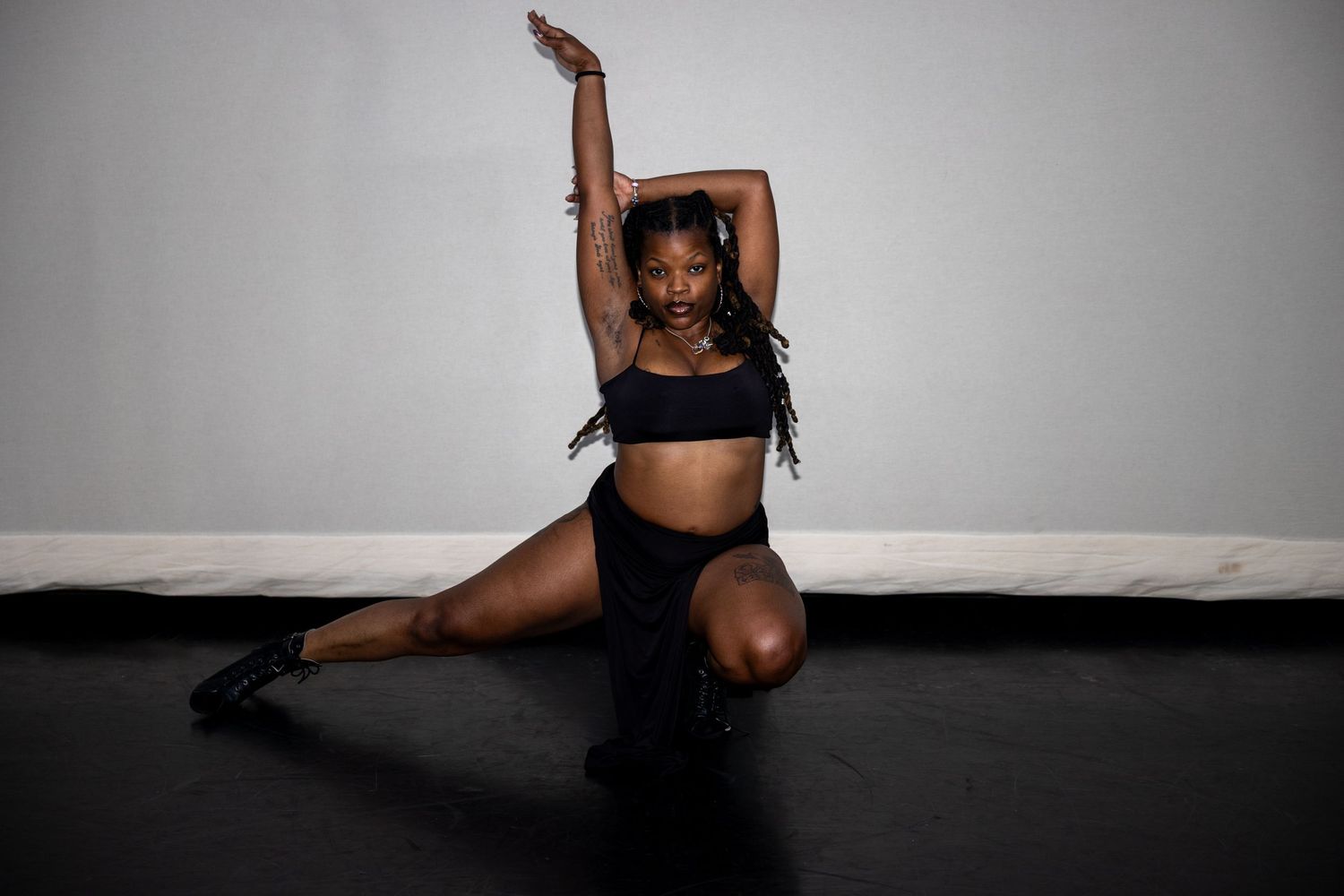 Dancer in black fringed crop top performing a dramatic floor pose with arm raised against white wall.