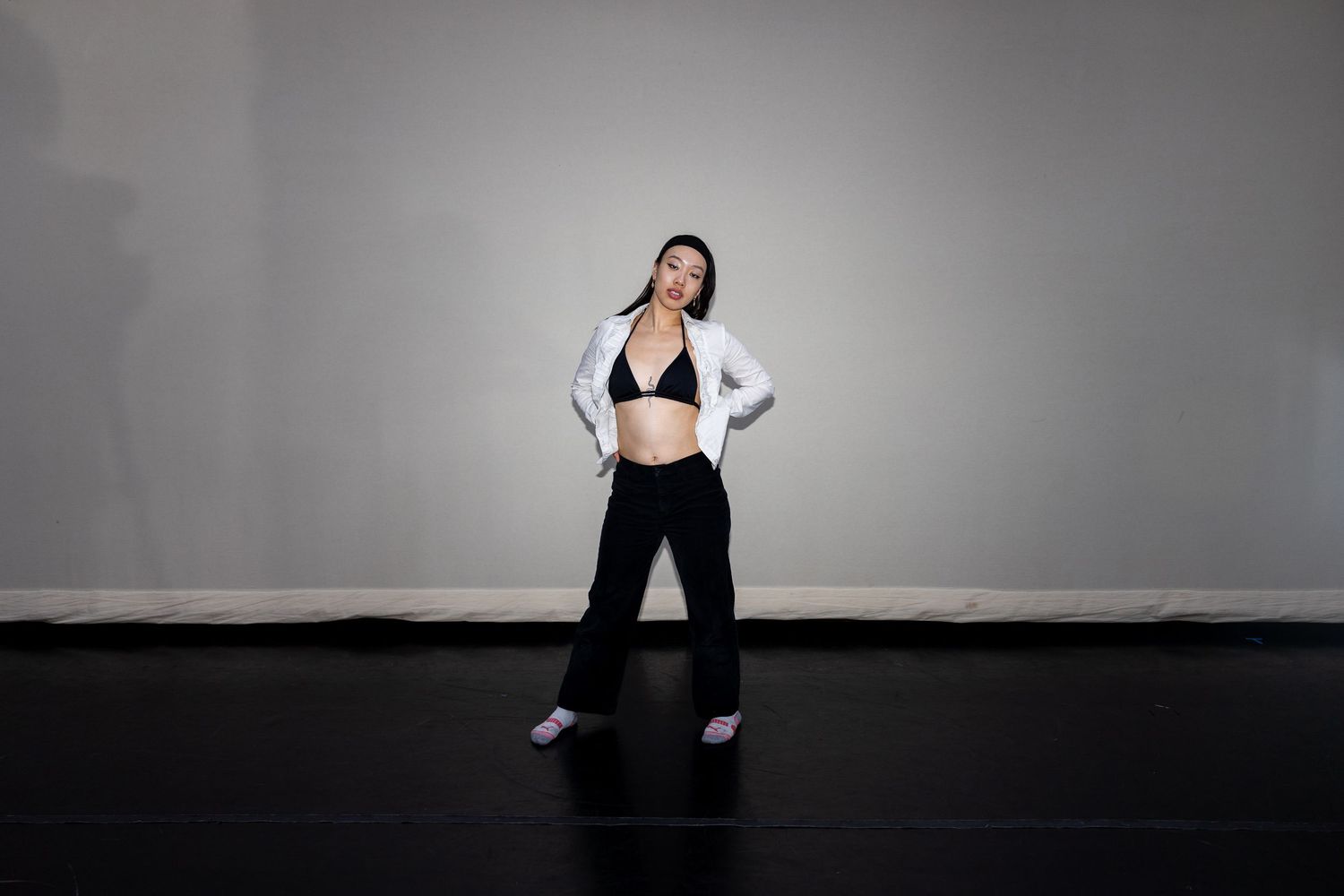 Woman in white blazer and black pants poses confidently on a dark stage floor against a gray wall.