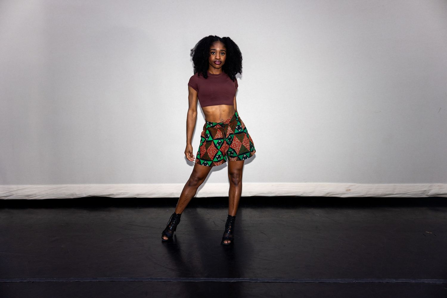 Black woman in colorful African print skirt and maroon crop top walking on a dark stage floor.