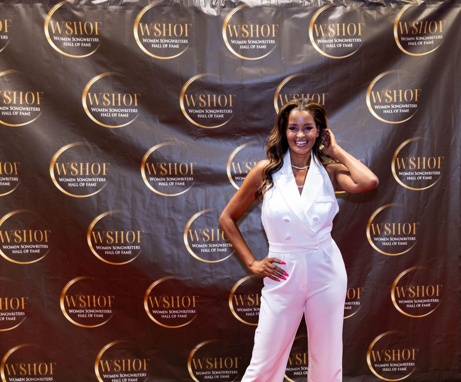 Woman in white jumpsuit poses at WSHOF Women Songwriters Hall of Fame event backdrop.